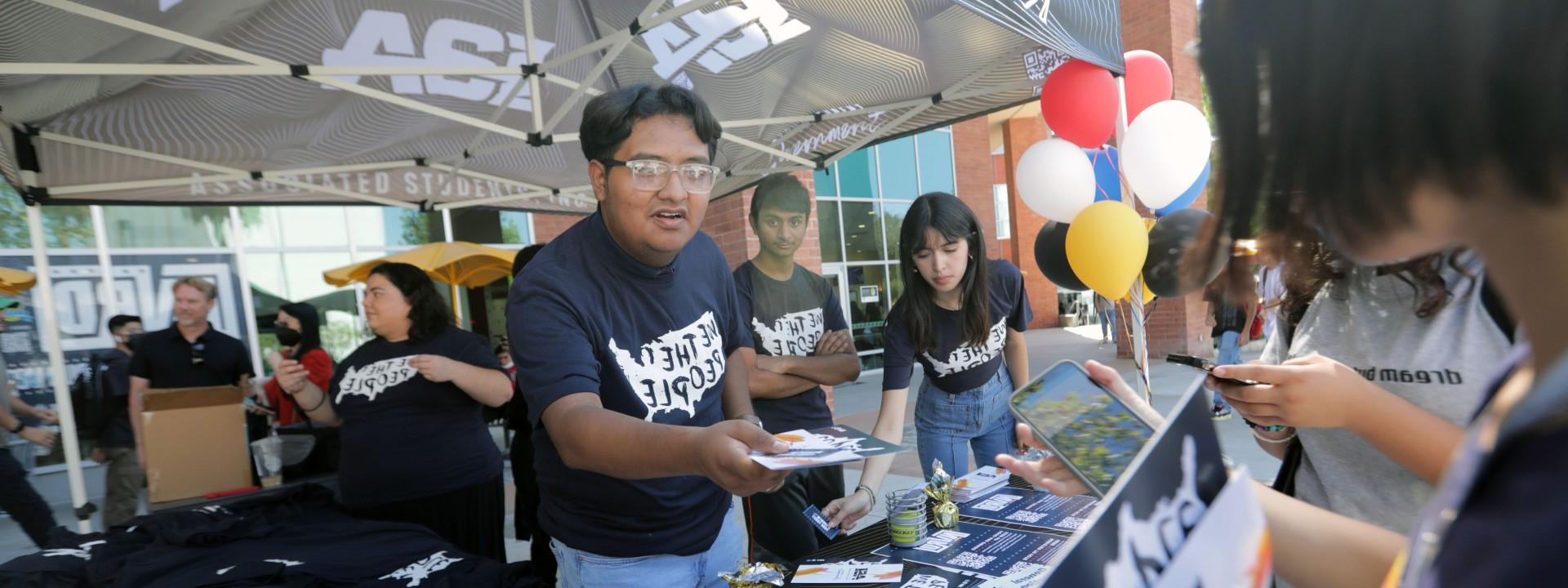 A student wearing a "We the People" t-shirt and standing under an Associated Students Inc., canopy hands students a brochure.